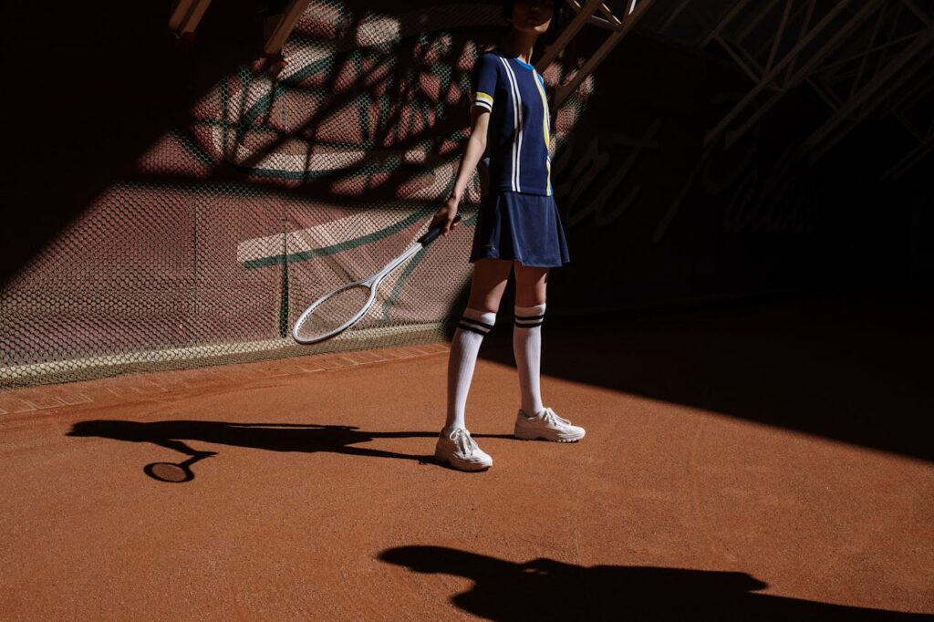 A young girl holding a tennis racket stands on a clay court, partially in shadow.